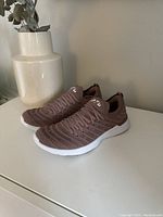 Side view of new brown APL running shoes with knit fabric and white soles, placed on a white surface next to a beige vase.