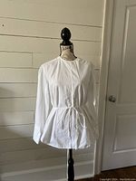 Front view of the white blouse displayed on a mannequin, showing long sleeves, round neckline, and waist tie.
