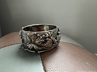 Silver cuff bracelet positioned on a surface showing raised floral and grape vine detailing, hinged clasp and attached safety chain.