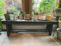Full frontal view of the black painted wooden bench with assorted plant pots on top, positioned near large windows with garden view.