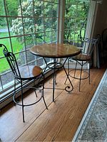 Full view of the bistro table set by window, showing round wooden table top and two black metal stools with wooden seats.
