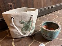 Both cement planters visible on a stone floor against wood paneling: the larger bag-shaped one with turtle detail and rope handles, and the smaller potted finish one with wear and crack.
