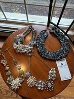 Four costume necklaces laid out on wooden surface near window: two chunky beaded necklaces and two silver-tone chain necklaces with floral clusters.