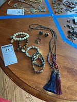 Photo showing two tassel necklaces, five beaded bracelets, one pair of earrings, and one pair of cuff links arranged on a wooden table.