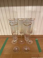 Six champagne flutes with heart-shaped, gold-rimmed tops displayed in two arrangements on a wooden surface against a paneled wall.