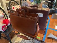 Two brown leather hard case briefcases, one with shoulder strap, placed on wooden chairs and table indoors.