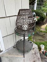 Front angled view of metal plant stand with wicker candle holder and solar candle on top, placed outdoors by a white house wall.