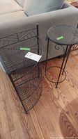Photo showing two black metal plant stands: one round stand with decorative top and one corner stand with three-tier wire shelves.