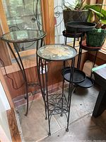 Three metal plant stands clustered together near window showing variation in tops: two with clear glass tops, one with decorative pressed flower top.