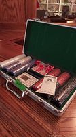 Open metal poker case showing stacks of poker chips in white, blue, and black along with five translucent red dice and two decks of playing cards in green and brown boxes.
