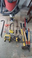 Overview of lot showing all tools on garage floor, including crowbars, hacksaw, caulking gun, work light, sander, shears, sledgehammer, and bolt cutters.