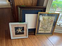 Three framed art pieces on wooden floor: white frame with starfish art, gold frame with floral print, and large dark frame with white mat, no glass.