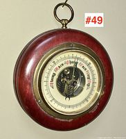 Front view of 1930s barometer showing dial, numbers and weather indications, with visible inner mechanism under glass, in a polished reddish wooden frame with a brass bezel and small hanging loop.