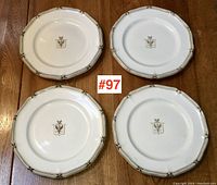 Four 19th century white plates with scalloped edges and gold trim, each displaying a central coat of arms, arranged on wood floor.