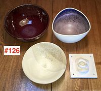 Overview of four pottery pieces on wooden table, showing red bowl, white bowl with speckled interior, cream crackle glaze bowl, and small square tile