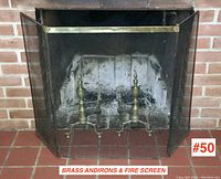 Wider view showing both brass andirons positioned inside fireplace with the brass-trimmed mesh firescreen in front.