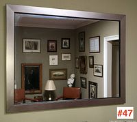 Front view of the large framed mirror hanging on a beige wall reflecting a room with framed artworks, a bust sculpture, and a lamp.