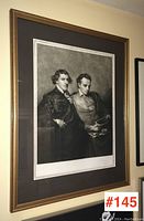 Framed signed black and white print hung on wall, showing two men in historical dress, one standing, one seated holding a book.