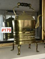 Front view of Victorian brass kettle on warming stand showing full height, wooden handle, hinged lid, body and ornate warming stand base with paw feet.