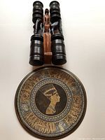 Photo showing two dark stained carved wooden candlestick holders, a carved wooden statue, and a round decorative plate with Cleopatra's profile and Egyptian motif design