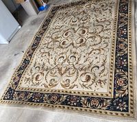 Full view of tan and black area rug showing intricate floral and scroll pattern with cream colored center and black floral border. Rug is on concrete floor in garage.