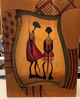 Framed carved wood panel artwork showing two stylized African figures in red and beige colors with geometric patterned border