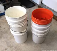 Side view of a stack of large white plastic buckets, five in total, with some showing wear and dirt, two on the right topped with orange-rimmed buckets.