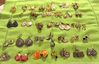 Top-down view of assorted silver and gold tone earrings laid out on a green cloth, showcasing a variety of styles including dangle, studs, and hoops, some with decorative detailing and colored accents.