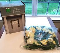 Photo showing the boxed 6-foot pre-lit Christmas garland with packaging labeled 'Happy Garland' and the spring wreath with yellow and white flowers still in plastic packaging.