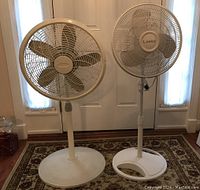 Two white floor fans positioned side by side, one Lasko brand with three blades and a rounded base, the other with five blades and a solid base. Both need cleaning.