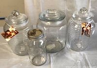 Four clear glass jars including one Mason jar with metal lid and three glass canisters with lids. Two ridged canisters have brand tags. Large hexagonal canister shows embossed branding.