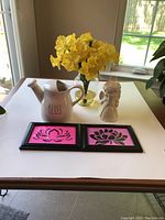 View of lot items including ceramic pitcher labeled 'Bliss', one angel figurine, framed lotus prints, and vase with faux daffodils on table near window.