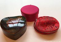 Set of three jewelry cases: a heart-shaped lacquered box, a round red fabric case, and a small red zippered pouch.