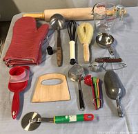 Photo of assorted kitchen tools including rolling pin, ladle, whisk, brush, measuring spoons, slicer, bench scraper, pizza cutter, oven mitts, and glass measuring cup