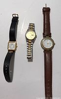 Three ladies wristwatches on a white background: rectangular gold-tone watch with black leather band, two-tone metal bracelet watch, and round gold-tone watch with brown leather band.