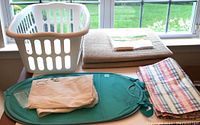 Laundry basket, pop-up hamper, two bath mats, plaid fabric items, and shower liners laid out on a surface with window background