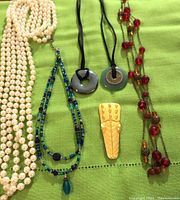 Top view of all necklaces and pin laid on green fabric background, showing long faux pearl necklace, red beaded necklace, blue-green beaded necklace, two medallion necklaces on black cords, and gold tone pin.