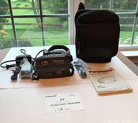 Photo showing Panasonic camcorder with power cord, AV cables, user manual, and soft carry case laid out on white table with window backdrop.