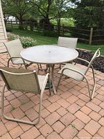 48-inch round glass-top patio table with four beige mesh sling chairs arranged around it on brick patio