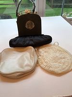 Four small handbags on a white surface by a window; includes a dark structured bag with gold-tone hardware, cream colored soft bag, woven round bag, and black clutch