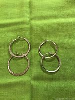 Two pairs of sterling silver hoop earrings displayed on green fabric; one pair is hammered texture and the other smooth.