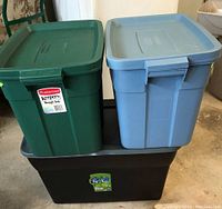 Three Rubbermaid Roughneck storage bins stacked. Two green 18 gallon bins on top side by side and one large black 30 gallon bin below with a blue lid on it.