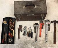 Vintage brown metal Stack-On toolbox with handle and two metal latches shown closed with assorted tools laid in front including pipe wrenches, pipe cutters, screwdrivers, hammer, and a tray with plumbing fittings.