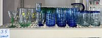 Full set of blue, green, and clear glassware arranged on shelf including pitchers, various glasses, and stemware.