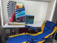 Full setup including blue and yellow collapsible chair with footrest, yellow Wiffle bat, white Wiffle ball, and pool box on a shelf with Boogie Boards behind.