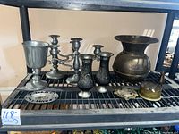 View showing two triple-arm pewter candle holders, a large pewter goblet, two small black floral decorative vases, a large brass vase, and multiple ashtrays including an oval pewter ashtray marked 'Paris France' and a brass Union Pacific spittoon.