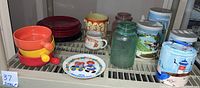 Photo showing handled ceramic soup bowls in orange, yellow, red, a Raggedy Ann and Andy 1976 Mother's Day plate, some red stoneware plates behind, and three canisters with farm scenes and a green glass canister.