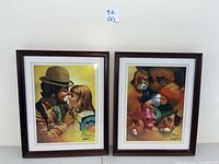 Two 15.5 x 19.5 inch wood-framed Chuck Oberstein clown prints side by side; one shows clown kissing a young girl, other shows multiple clown faces.