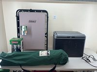 Wide view of all four items on display: cot folded against the wall, green bag likely for the chair, lantern boxed, and plug-in cooler on white table.