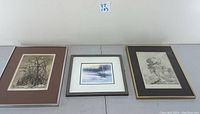 Three framed and matted artworks including two silver/wood framed prints and one gold framed etching with broken glass on the right.